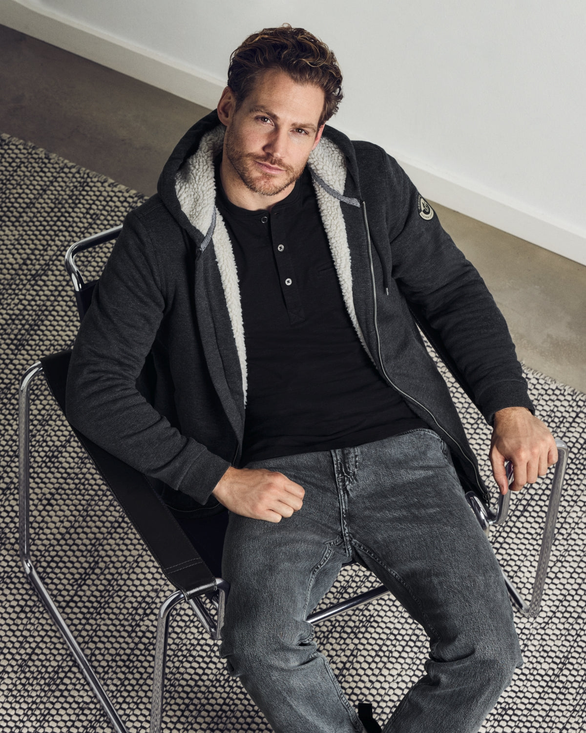 Ein Mann, der ein schwarzes Hemd, eine dunkelgraue Kapuzenjacke mit Reißverschluss und hellem, flauschigem Innenfutter sowie eine dunkelgraue Jeans trägt.