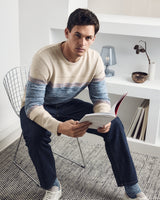 Ein Mann trägt einen beige-blau gestreiften Pullover, dunkelblaue Jeans und weiße Turnschuhe und hält ein aufgeschlagenes Buch in der Hand.