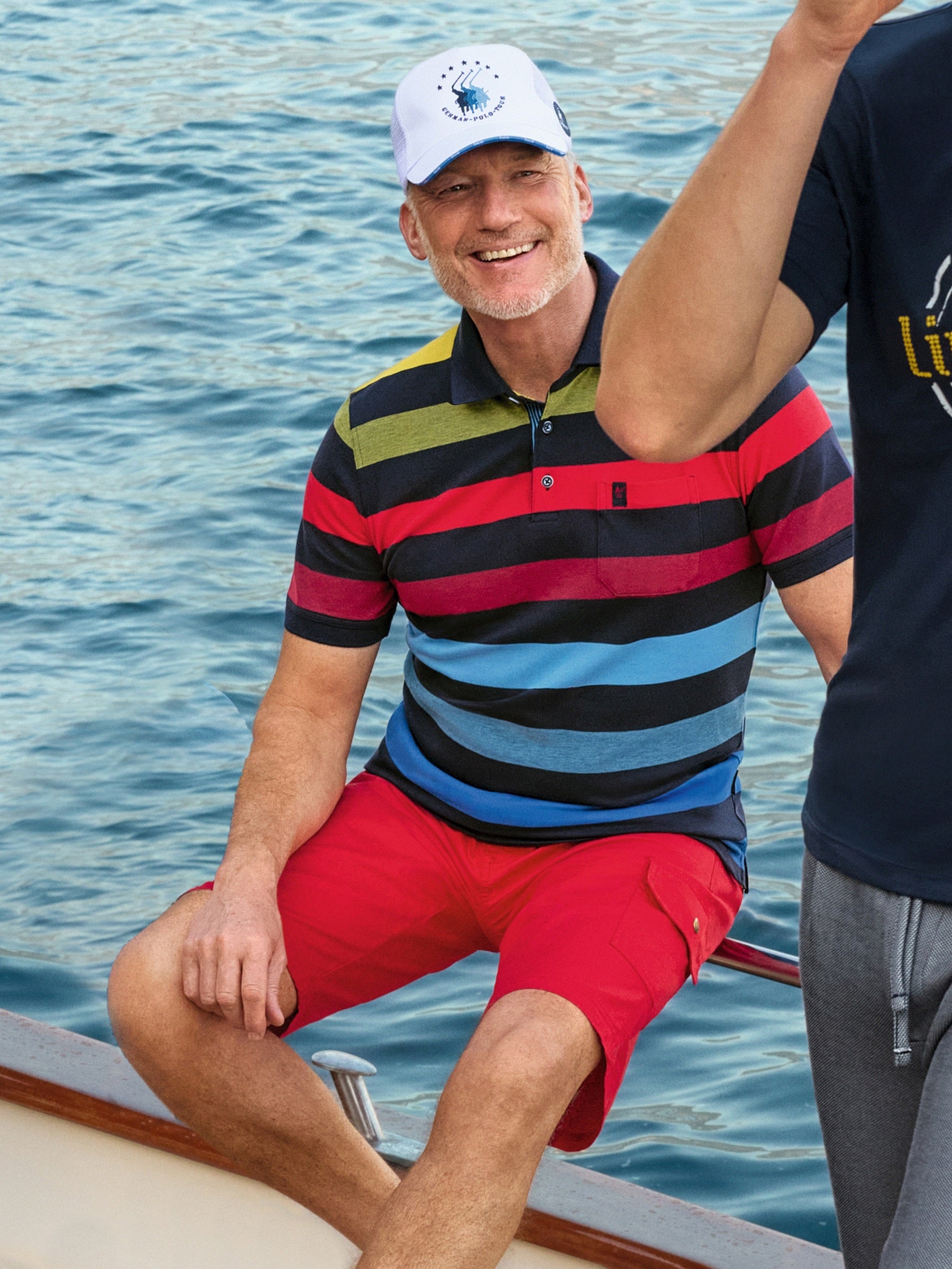 Mann in weißer Pikee-Cap mit blauem Logo, bunt gestreiftem Blockringel-Polo und roten Shorts
