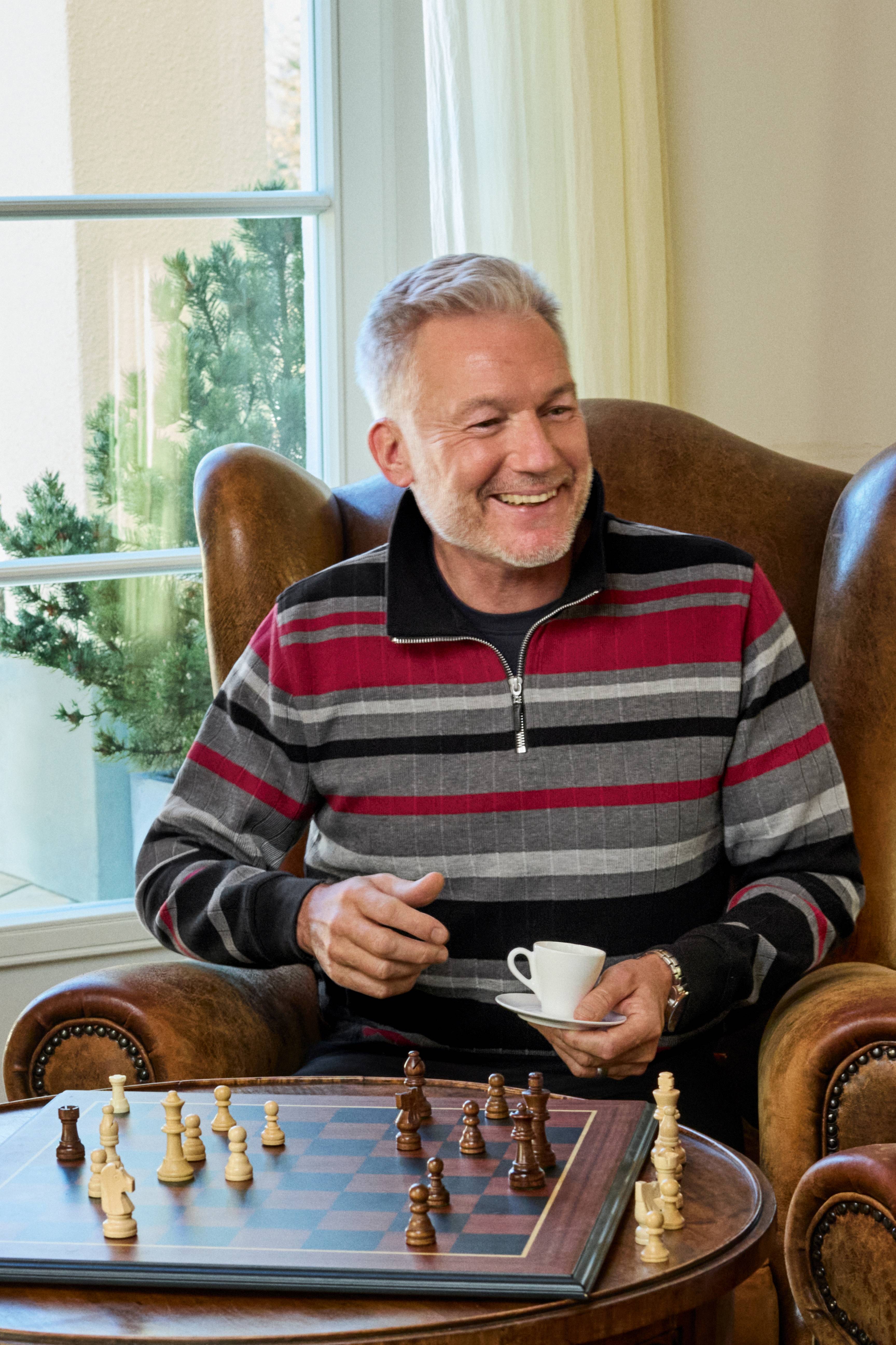 Ein mann sitzt lächelnd mit einem weißen keramikkaffeebecher in einem rot grau schwarz gestreiften troyersweatshirt