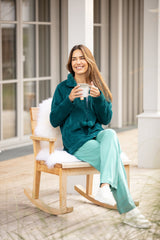 Eine Frau in einer blaugrünen Button-up-Jacke und einer hellgrünen Hose mit einer Tasse in der Hand sitzt auf einem hölzernen Schaukelstuhl mit einer weißen Plüschdecke.