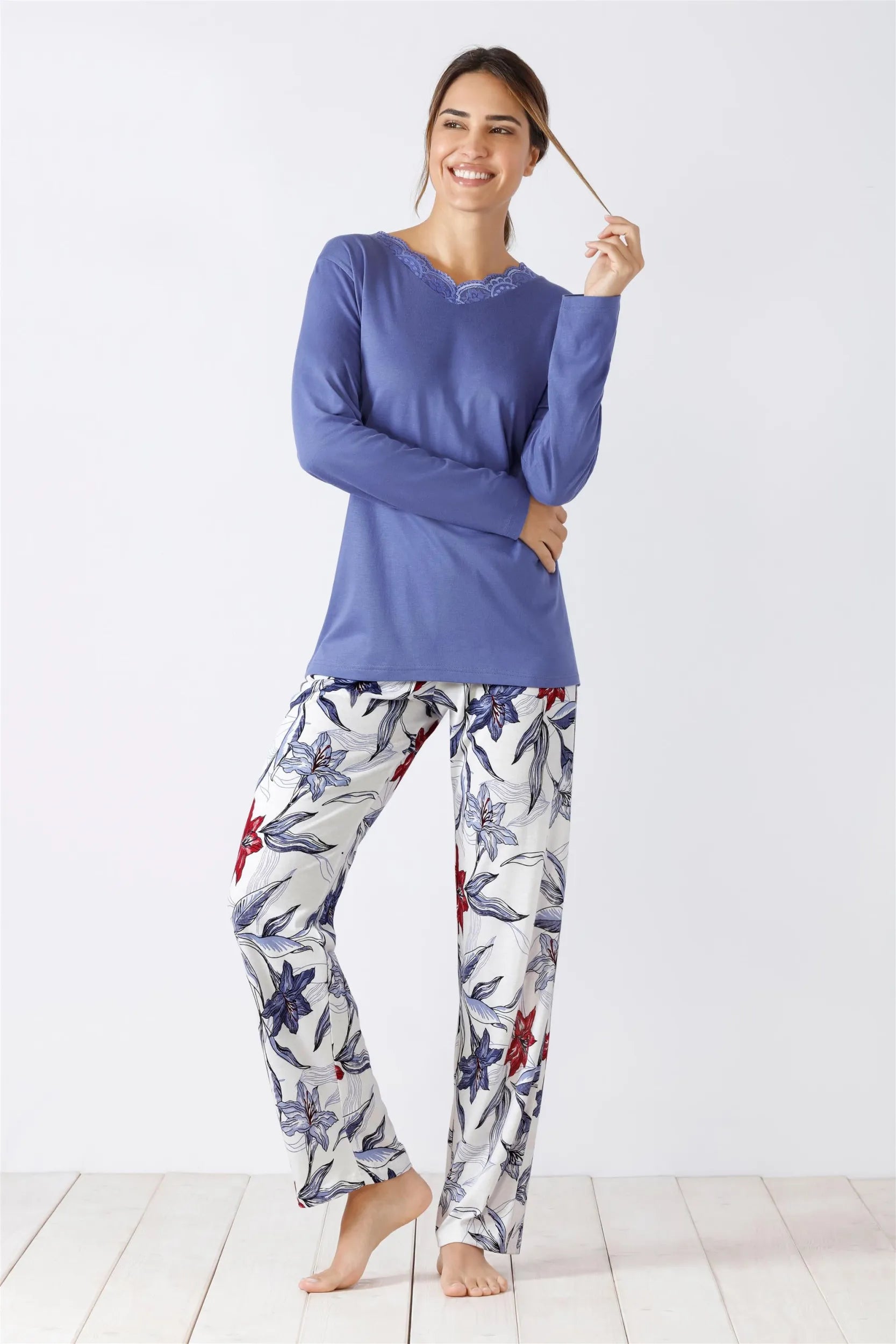Eine Frau modelliert ein langärmeliges, violettfarbenes Oberteil mit dezentem V-Ausschnitt und eine weiße Pyjamahose mit einem Blumenmuster aus blauen und roten Blüten.