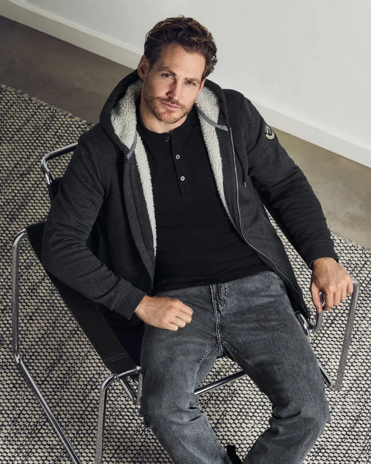 Ein Mann, der ein schwarzes Hemd, eine dunkelgraue Kapuzenjacke mit Reißverschluss und hellem, flauschigem Innenfutter sowie eine dunkelgraue Jeans trägt.