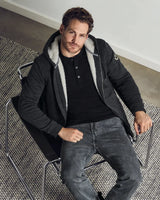 Ein Mann, der ein schwarzes Hemd, eine dunkelgraue Kapuzenjacke mit Reißverschluss und hellem, flauschigem Innenfutter sowie eine dunkelgraue Jeans trägt.