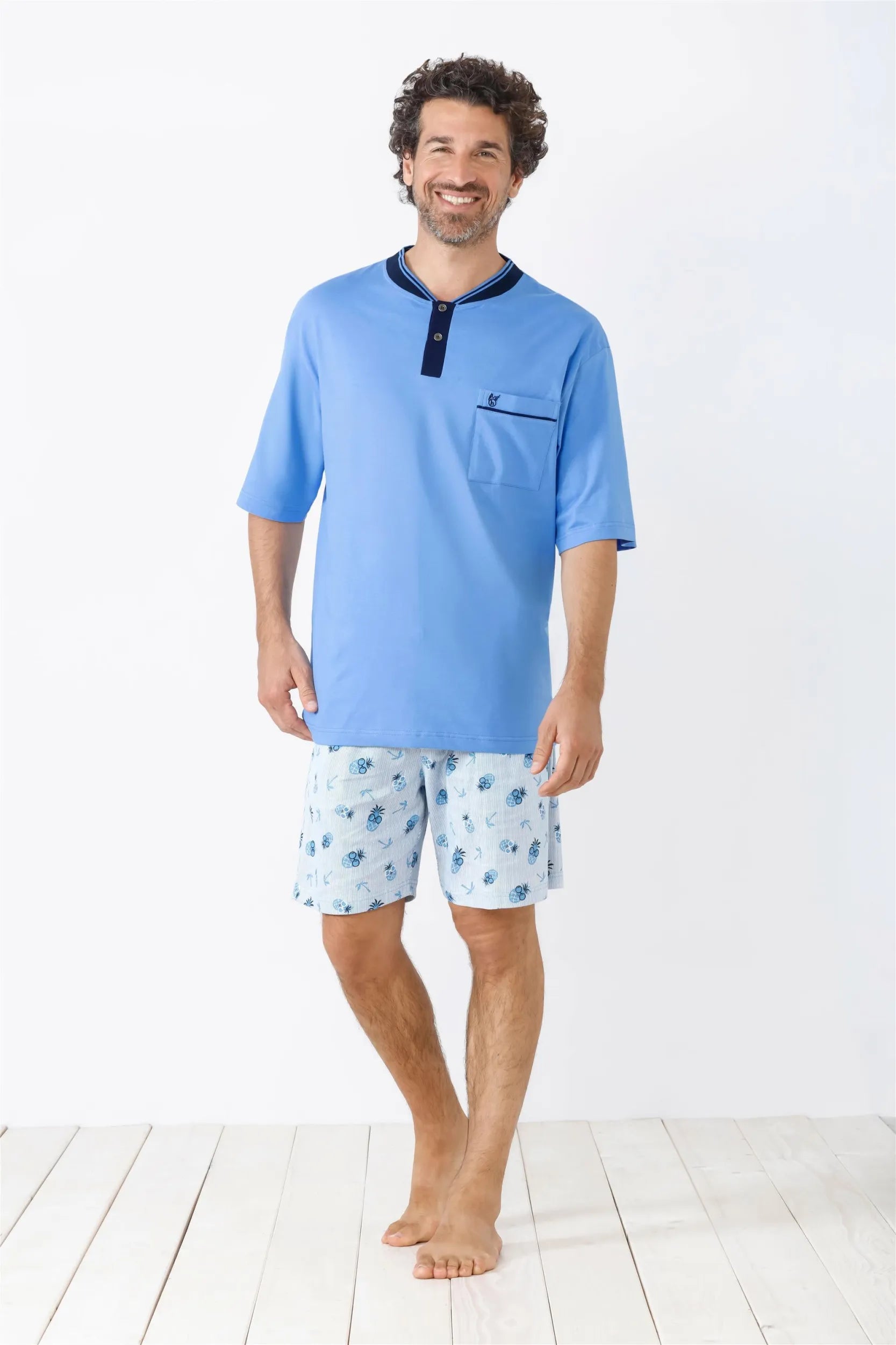 Hellblaues kurzärmeliges Herren-Pyjama-Oberteil mit dunkelblauer Knopfleiste und Tasche, kombiniert mit hellblau gemusterten Pyjama-Shorts.