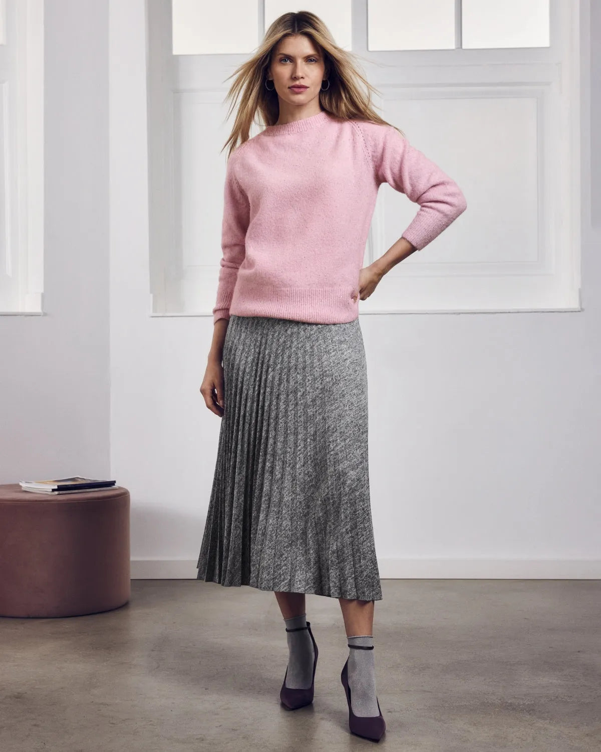 Eine Frau trägt einen rosafarbenen langärmeligen Pullover und einen grauen plissierten Midirock.