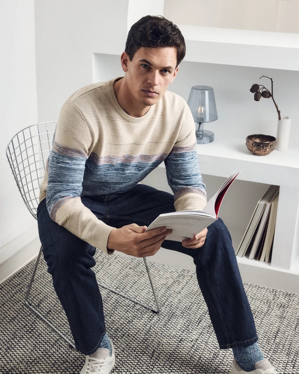 Ein Mann trägt einen beige-blau gestreiften Pullover, dunkelblaue Jeans und weiße Turnschuhe und hält ein aufgeschlagenes Buch in der Hand.